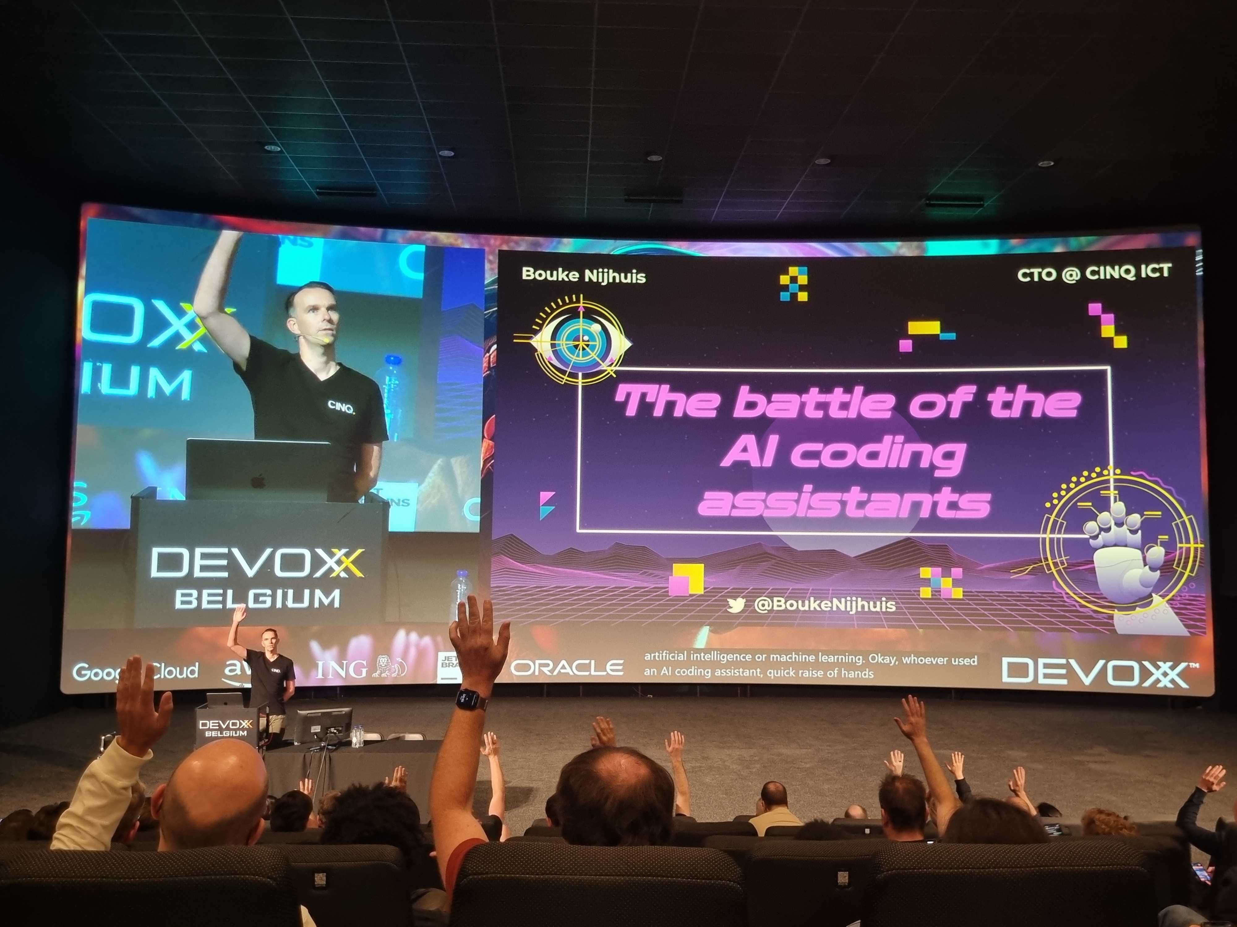 A presenter with one arm raised behind podium with screen titling Battle of the AI Coding Assistants.